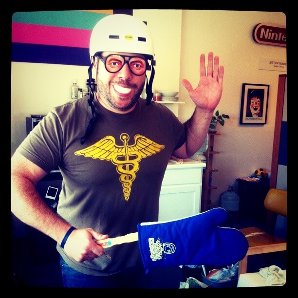 Mike Roush waving, wearing a helmet, glasses with fake eyes, and a mouth protector, holding an oven mitt on a stick.